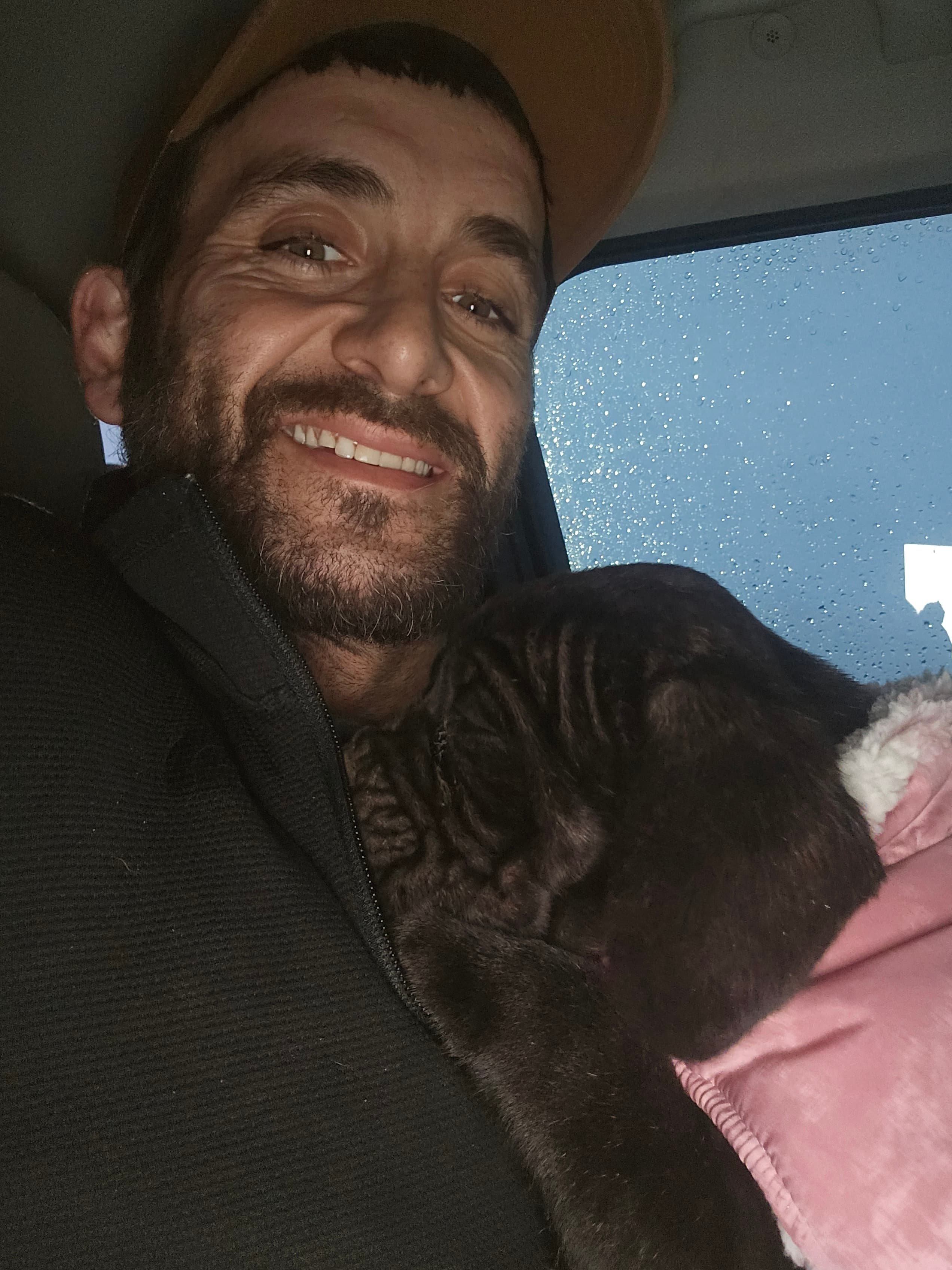 Smiling bearded man holds a sleeping wrinkly puppy wrapped in a pink blanket.